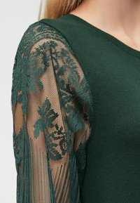 Πράσινο πλεχτό μπλουζάκι με μακριά, διάφανη δαντέλα στα μανίκια που παρουσιάζει λουλουδένια μοτίβα και υφασμάτινες λεπτομέρειες. Στρογγυλή λαιμόκοψη, εφαρμοστή γραμμή.