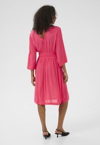 Robe rose avec des manches cloche trois-quarts, taille froncée et dos en forme de clé. Tissu transparent avec une texture légère et fluide.
