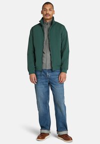 Grüne, leichte Jacke mit Reißverschluss auf der Vorderseite, kombiniert mit einer grauen Fleece-Innenlage, blauen Jeans und braunen Stiefeln. Lässiges Outfit für kühles Wetter.