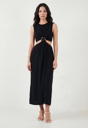 Kvinde med bølget mørkt hår iført en ærmeløs sort kjole med udskæringer i siden og en guldlåsdetalje, kombineret med beige ankelstroppestiletter.