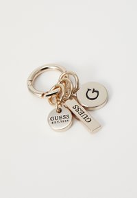 Guldtonet nøglering med runde og rektangulære mærker; et mærke har "GUESS EST.1981" og et andet viser et stiliseret "G" logo.