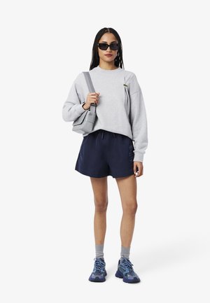 Grauer Sweatshirt, marineblaue Shorts, graue Socken und blaue Sportschuhe. Sonnenbrille und graue Tasche. Auffällig durch ihr lässiges Design.