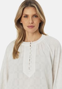 Hvid blouse med et struktureret ternet mønster, rund halsudskæring og en stolpe med fem knapper. Blødt, let stof.