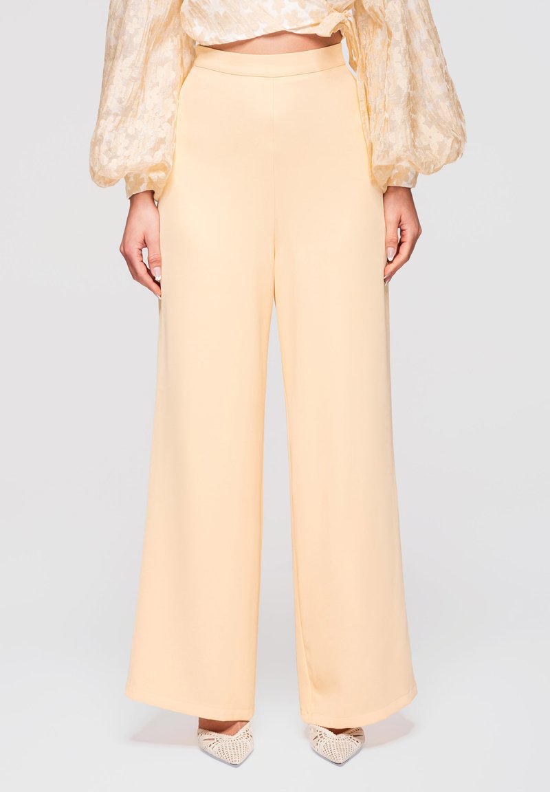 Femme portant un pantalon beige à taille haute et jambes larges, associé à un chemisier transparent à manches longues aux motifs floraux et des chaussures blanches tissées à bout pointu.