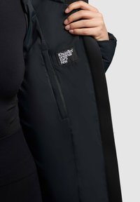Černá prošívaná bunda s elegantním designem, která má výraznou visačku loga na vnitřní podšívce a hladkou texturu.