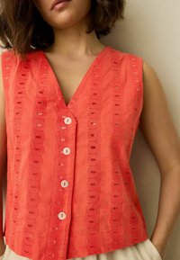 Haut sans manches corail avec un col en V profond, orné de motifs ajourés complexes et six boutons blancs devant.