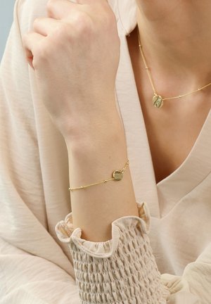Gold bracelet with a smooth, rounded charm, featuring a fine chain and small beads. Worn on a wrist with textured sleeve detail.