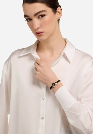 Klassische Armbanduhr mit einem schwarzen Lederarmband, runder Gehäuse in Goldton und schwarzem Ziffernblatt. Modell trägt ein weißes Hemd mit Knöpfen.