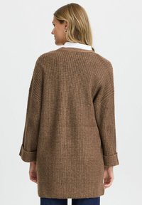 Brauner, gestrickter, übergroßer Cardigan mit einem strukturierten Muster, langen Ärmeln und umgeschlagenen Bündchen, getragen über einem weißen Hemd mit Kragen.