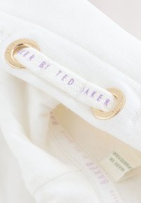 Gros plan sur un tissu blanc avec des œillets métalliques dorés et une étiquette tissée portant la mention « Baker by Ted Baker » en texte violet clair.
