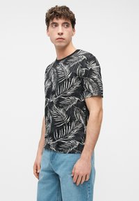 Νεαρός άνδρας που φοράει μαύρο T-shirt με λευκό σχέδιο φύλλου και ανοιχτό μπλε τζιν, κοιτάζει προς τα αριστερά του, με λευκό απλό φόντο.