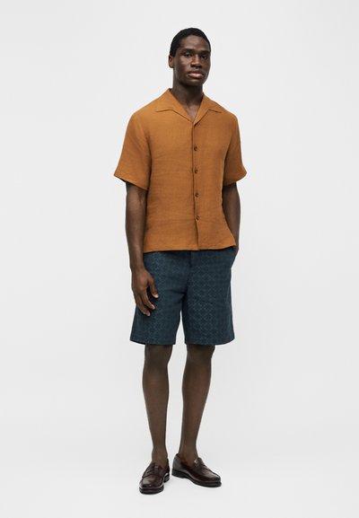 Άνδρας στέκεται φορώντας κοντομάνικο καφέ πουκάμισο με κουμπιά, σκούρα μπλε σορτς με σχέδια και σκουρό καφέ μοκασίνια, πάνω σε απλό φόντο.
