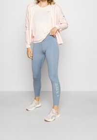 Giacca leggera rosa chiaro sopra una T-shirt Nike grigia chiara, abbinata a leggins blu con logo bianco e dettagli bianchi, e sneakers chiare.