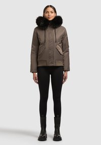 Ruskea toppatakki, jossa on musta turkisvuorinen huppu, etutaskut ja kiristysnyörit, yhdistetty mustiin trikoihin ja nilkkureihin.