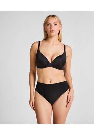 Blondynka ubrana w czarny podszywany biustonosz i czarne majtki z wysokim stanem, stojąca na jednolitym szarym tle.