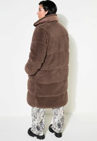 Cappotto imbottito marrone in pelliccia finta, morbido e soffice, con colletto alto, abbinato a pantaloni a stampa serpente texturizzati e stivali neri chunky.