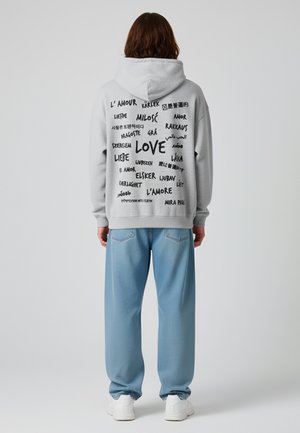 Sweat à capuche gris avec plusieurs phrases "amour" imprimées en plusieurs langues au dos, associé à un pantalon ample bleu clair.