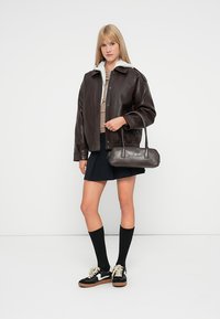 Veste en cuir marron avec un col gris, jupe noire et chaussettes montantes noires. Tenant un petit sac à main sombre, portant des baskets noires.