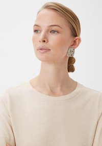 Srebrne, teksturowane kolczyki w kształcie kwadratu noszone przez modelkę. Góra zdobiona falistymi wzorami i polerowanym wykończeniem. Prosta beżowa bluzka wyeksponowana.