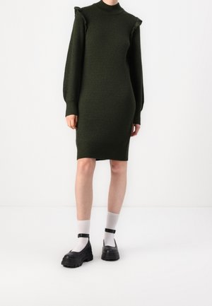 Robe en maille vert foncé à silhouette ajustée, texture côtelée, col montant et épaules volantées ; associée à des chaussures noires épaisses et des chaussettes blanches.