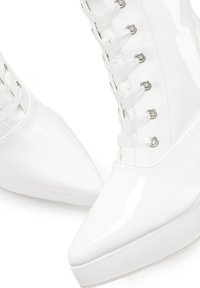 White patent leather platform boots with a pointed toe, featuring silver eyelets and satin laces, highlighting a smooth, glossy texture.