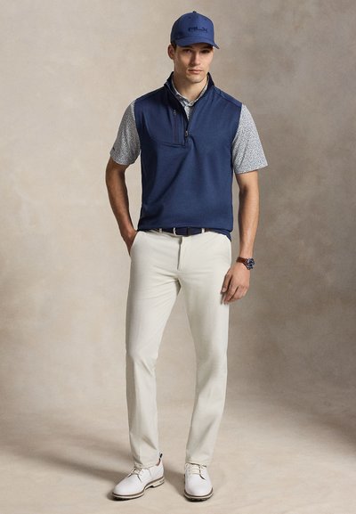 Homme portant une casquette bleu marine et un gilet par-dessus une chemise à manches courtes à motifs, pantalon beige, chaussures blanches, debout avec une main dans la poche.