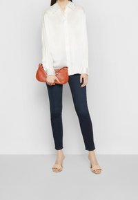 Camisa blanca de satén con botones, jeans ajustados azul oscuro, bolso de coral con acento metálico y tacones abiertos beige. Texturas suaves y diseño ajustado.