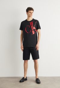 T-shirt en coton noir avec un grand logo CK rouge, associé à un short noir et des chaussures noires à enfiler. Design simple et coupe décontractée.