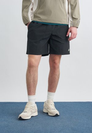 Mand iført sorte sportsshorts, beige sneakers, hvide sokker og en beige jakke, der står på blåt tæppe.