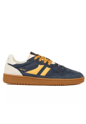 Sneaker basso in camoscio blu navy, strisce gialle, lacci beige, tallone bianco e suola in gomma marrone, che mostra il dettaglio "1989" sul lato.