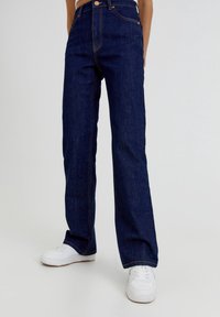 Jeans de mezclilla de tiro alto en un tono azul oscuro, con corte recto, costuras en contraste en color naranja y diseño clásico de cinco bolsillos, combinados con zapatillas blancas.