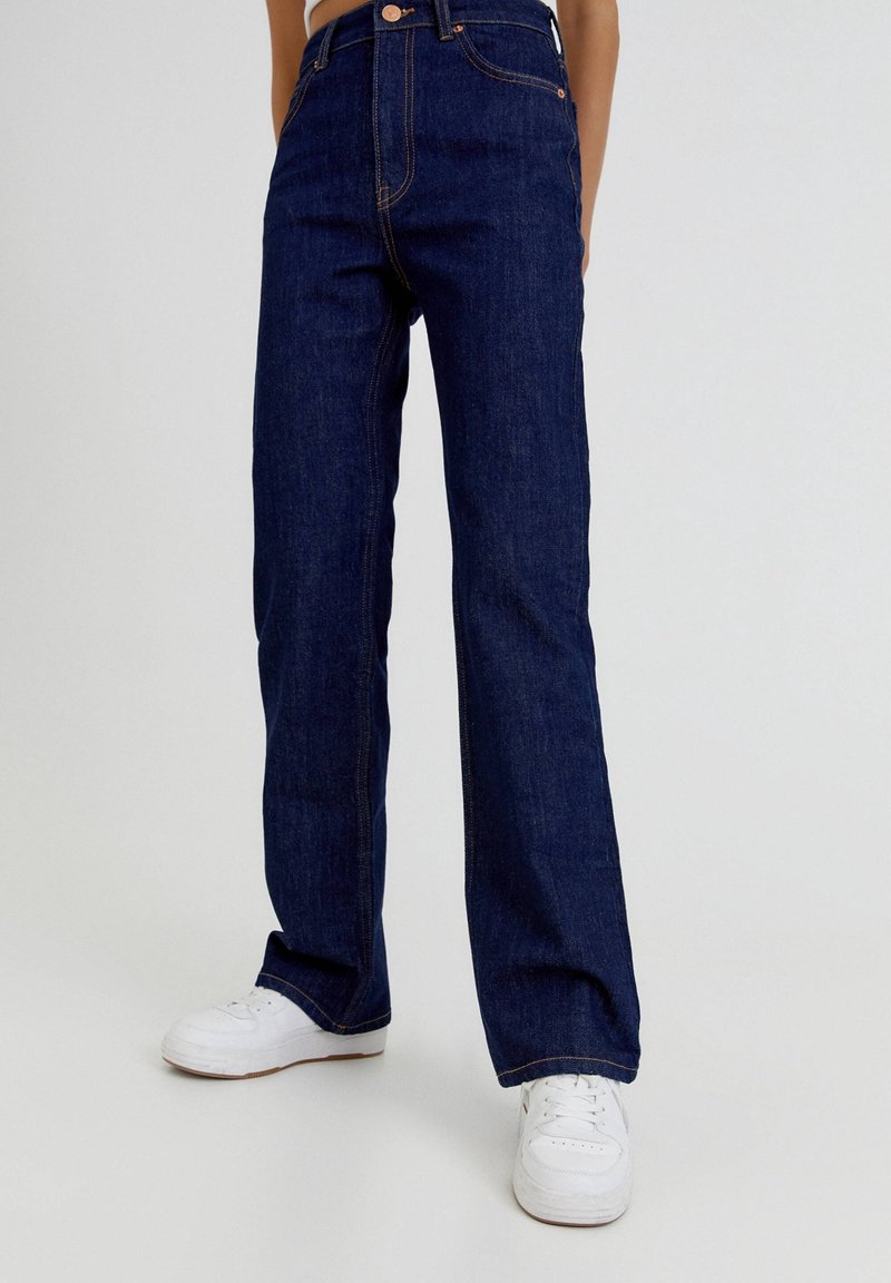 Jeans de mezclilla de tiro alto en un tono azul oscuro, con corte recto, costuras en contraste en color naranja y diseño clásico de cinco bolsillos, combinados con zapatillas blancas.