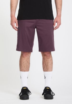 Homme portant un short violet foncé, un t-shirt noir, des chaussettes blanches montant jusqu'au milieu du mollet et des baskets noires, debout sur un fond blanc.