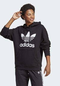 Černá mikina s kapucí a bílým logem Adidas a vzorem tří pruhů. Má přední kapsu a volný střih.