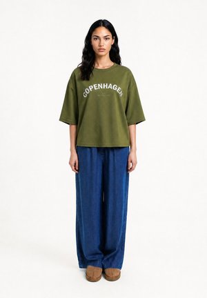 Frau mit langen dunklen Haaren, die ein olivgrünes oversized T-Shirt mit dem Text "Copenhagen", weite blaue Jeans und braune Schuhe trägt und frontal steht.