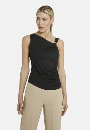 Vrouw die een zwart asymmetrisch mouwloos topje met gedrapeerd detail draagt en beige broek met hoge taille, lichtjes opzij kijkend.