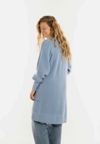 Vrouw met lang, krullend haar die glimlacht, in een lichtblauwe gebreide cardigan en blauwe spijkerbroek, zijdelings staand tegen een effen achtergrond.