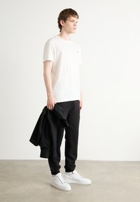 Λευκό βαμβακερό t-shirt, μαύρα αθλητικά παντελόνια και λευκά αθλητικά παπούτσια. Το μοντέλο κρατά μια μαύρη ζακέτα. Λείος υφασμάτινος πηλός, άνετη εφαρμογή, μινιμαλιστικός σχεδιασμός.