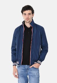 Giacca bomber blu navy con zip frontale, dettagli bianchi e rossi, polsini elasticizzati e finitura texture, abbinata a un polo nera e jeans strappati.
