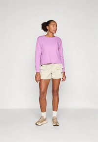 Γυναίκα που φοράει ένα μακρυμάνικο πουκάμισο λεβάντας, μπεζ cargo σορτς, λευκές κάλτσες και μπεζ αθλητικά παπούτσια, που στέκεται μπροστά από ένα απλό φωτεινό φόντο.