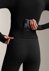 Černý sportovní top s dlouhým rukávem a přiléhavé legíny, které mají nenápadnou kapsu na telefon na zadní straně, vyrobené z elastického a hladkého materiálu.