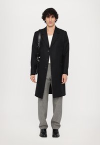 Schwarzer langer Mantel mit Reverskragen und Knopfverschluss, kombiniert mit grauen karierten Hosen und schwarzen Schuhen. Das Modell hält eine kleine Tasche.