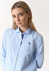 Giovane donna con capelli biondi che indossa una camicia a righe azzurre e bianche con bottoni e un piccolo logo ricamato blu navy sul petto.