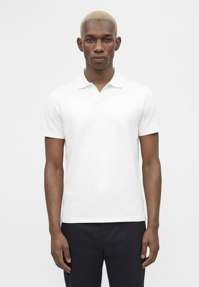 Homme aux cheveux courts décolorés portant un polo blanc uni à manches courtes et un pantalon noir, debout devant un fond blanc.