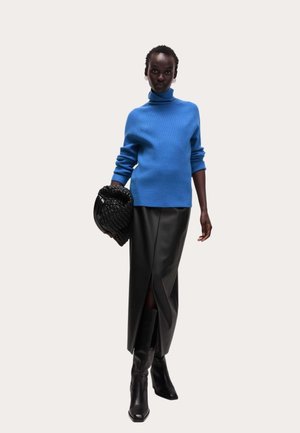 Pull à col roulé en maille bleue, jupe midi en cuir noir fendillée, tenant un sac à main tissé noir, associé à des bottes à talons noires.