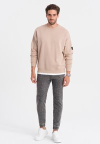 Ljusbeige sweatshirt med lång ärm, rund halsringning och svart patch på vänster arm, tillsammans med grå texturerade joggers och vita sneakers.
