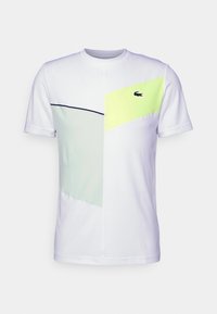 Vit T-shirt med gröna och gula geometriska paneler, korta ärmar, rund halsringning och en liten Lacoste-logotyp på bröstet.