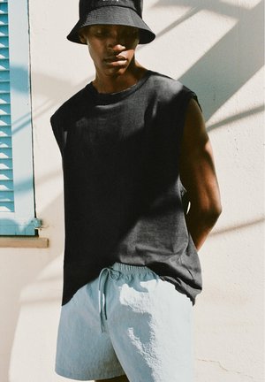 Un tânăr purtând o pălărie neagră tip bucket, un tricou fără mâneci negru și pantaloni scurți bleu stă lângă un perete alb cu o fereastră cu obloane albastre.