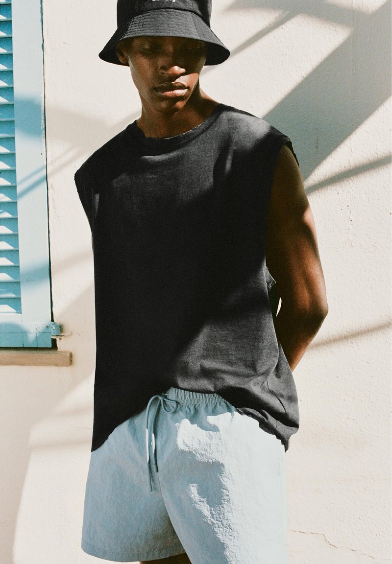 Jeune homme portant un chapeau bob noir, un débardeur noir et un short bleu clair, debout près d'un mur blanc avec une fenêtre à volets bleus.