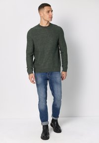 Vihreä teksturoitu neule pyöreällä pääntiellä, yhdistettynä sinisiin kapealahkeisiin farkkuihin ja mustiin nilkkureihin. Asu on istuvuudeltaan muotoiltu ja värimaailmaltaan neutraali.